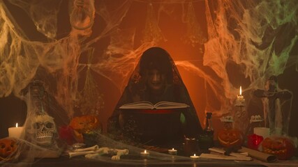 Witch in black clothes reading spell book in scary kitchen with pumpkins and human bones.