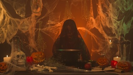 A witch in black clothing is cooking in a cauldron in a scary kitchen with pumpkins and a human skull and bones.