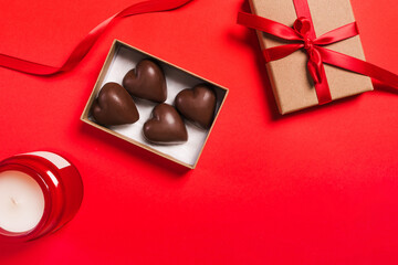 A gift box containing heart-shaped chocolates decorated with a red ribbon on a red background with a candle