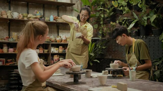 Medium long shot of two diverse kids listening to female Asian teacher guiding them during pottery masterclass in workshop