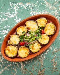 Clay plate is presenting round mushrooms topped with melted cheese and garnished with fresh greens