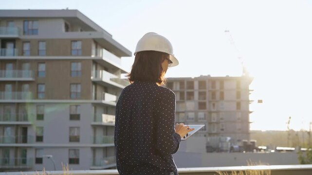 Woman architect wearing a hard hat is taking notes with a tablet computer while inspecting a building construction site at sunset