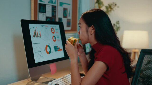 Young businesswoman is eating a sandwich while working on her computer. She is sitting at her desk in her office and is looking at a graph on the screen