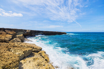 Rocky coastline with waves crashing against the shore, bright blue Caribbean Sea under a partly cloudy sky in Aruba.