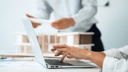Close-up of young beautiful professional architect hands using laptop with blueprint and architectural document placed on table while male coworker inspects house model. Focus on hand. Immaculate.