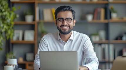 A smiling man in a white shirt working on a laptop in a modern office with books and plants in the background.