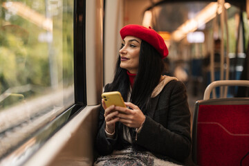 Woman wearing a red beret and coat, using her phone while enjoying a train ride, looking out the...