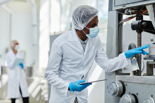 African American male process technician in lab coat looking inside manufacturing machine controlling quality and safety of production process on shop floor of pharmaceutical factory, copy space