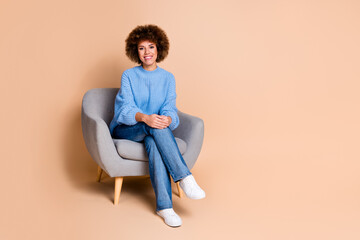 Full size photo of positive girl dressed knit sweater jeans sit on armchair near discount empty space isolated on pastel color background