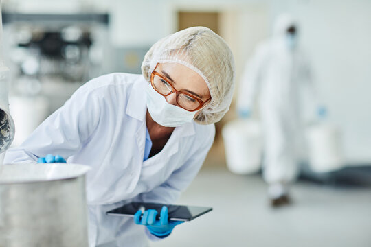 Female manufacturing supervisor in protective mask and glasses overseeing equipment while controlling production process and documenting results in digital tablet at pharmaceutical plant, copy space