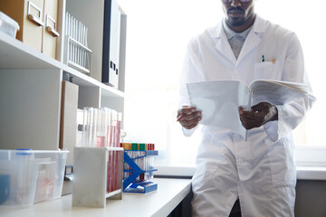 Fototapeta premium Cropped shot of African American male lab chemist reading book next to window performing scientific research in pharmaceutical laboratory, copy space