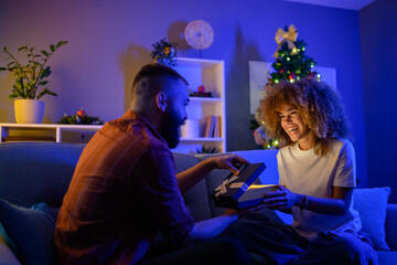 Young man giving christmas present to girlfriend sitting on sofa