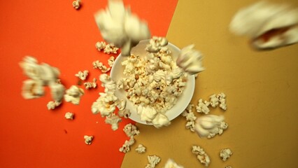 Top down view of flying popcorn dropping at white bowl with yellow and orange background. Super slow motion shot of piles of fluffy salted popcorn falling in to the bowl. Macrography. Comestible.