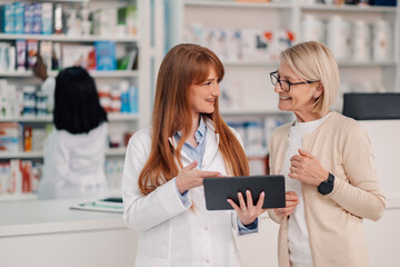 Female pharmacist using digital tablet while helping senior customer in pharmacy