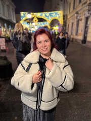 Millennial woman standing outdoors at Christmas night, wrapped in a cozy shearling jacket. the city lights in the background create a warm and Christmas holidays atmosphere, enhancing the peaceful and