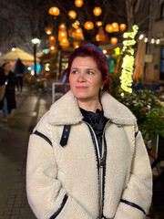 Millennial woman standing outdoors at Christmas night, wrapped in a cozy shearling jacket. the city lights in the background create a warm and Christmas holidays atmosphere, enhancing the peaceful and