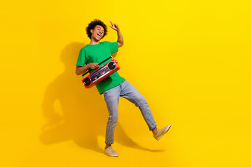 Fototapeta premium Full length photo of handsome good mood guy wear green t-shirt showing v-sign enjoy boom box songs emtpy space isolated yellow color background