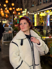 Fototapeta premium Millennial woman standing outdoors at Christmas night, wrapped in a cozy shearling jacket. the city lights in the background create a warm and Christmas holidays atmosphere, enhancing the peaceful and