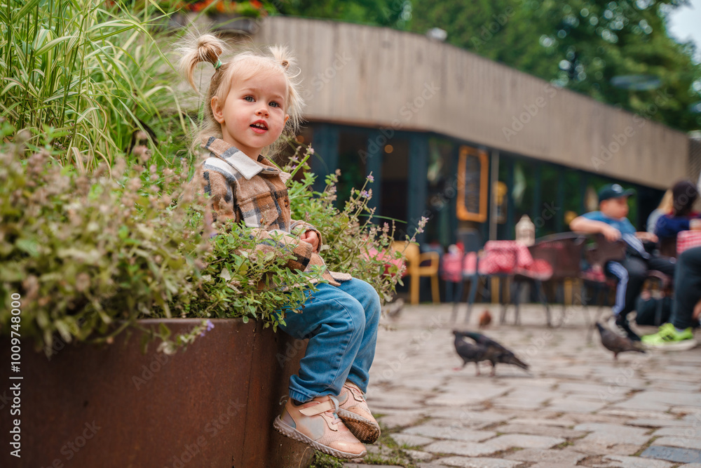 Wall mural a cute toddler kid girl in a plaid shirt sits on the veranda of a european outdoor cafe in summer.