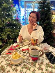 Millennial woman enjoys festive Christmas meal at decorated table, surrounded by Christmas trees and twinkling lights, capturing warmth and joy of holiday season. Joyful expression and cozy setting