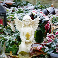 a small white figure of an angel with a cross in folded hands on a grave with plants covered in...