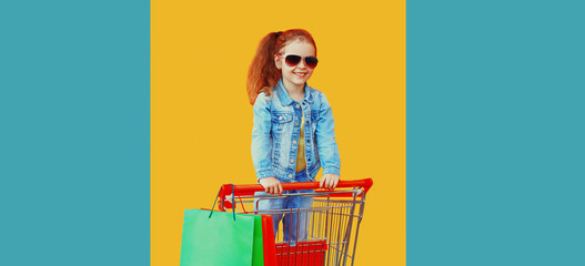 Happy smiling little girl child with trolley cart, shopping bags on colorful yellow background