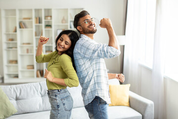 Joyful indian couple dancing in living room interior, having fun and singing, husband and wife...