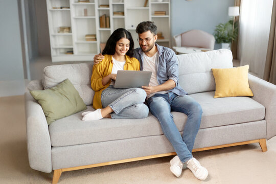 Planning vacation. Happy young indian couple searching tour online, using laptop computer, sitting on couch in cozy living room interior at home, free space