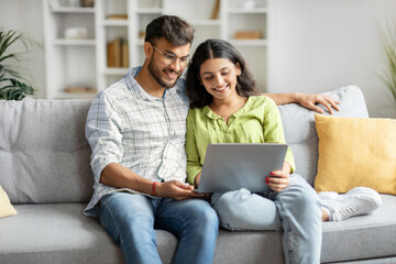 Loving happy indian couple sitting on couch at home, embracing, using laptop computer, young man and woman watching movie online, copy space