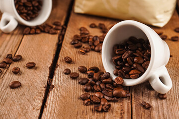 Roasted beans for mass market sale. Spilled coffee beans from two white ceramic cups on brown wooden background. Coffee shop, caffeine, roast concept.