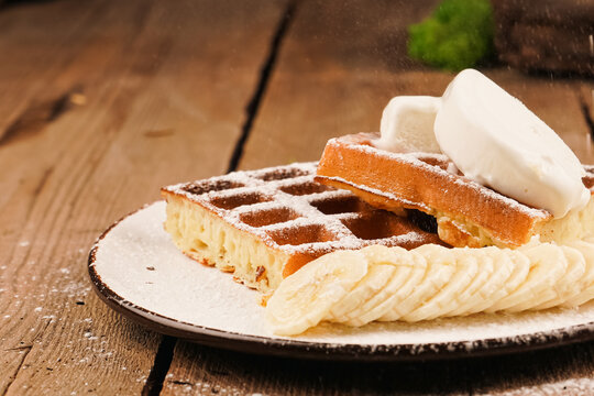 Belgian waffles on wooden table. Belgian waffles on a white plate. Topped powdered sugar. Waffles on plate. Ice-cream topping. Cooking process. Adding banana on waffle. Homemade simple pastries.