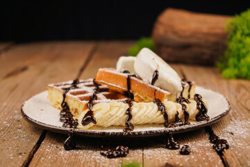 Sweet dessert waffles. Belgian waffles with banana, ice cream and chocolate on top. Delicious dessert for tea time. Served with ice cream. Plate of waffles with ice cream and banana on wooden table.