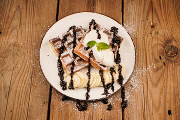 Served with ice cream. Belgian waffles with banana, ice cream and chocolate on top. Sweet dessert waffles. Delicious dessert for tea time. Mint on top of dessert. Cooking process. Top view.