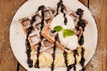 Belgian waffles with banana, ice cream and chocolate on top. Sweet dessert waffles. Served with ice cream. Delicious dessert for tea time. Mint on top of dessert. Cooking process. Top view.