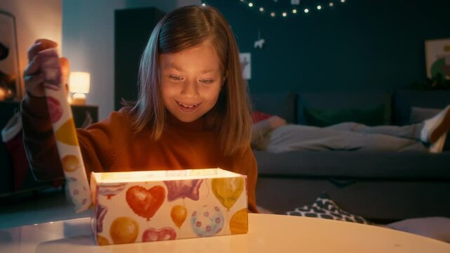 Chest up shot of little naughty girl in brown sweater looking at sleeping granddad and furtively opening Christmas present shining from inside and getting surprised in decorated living room
