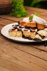 Delicious dessert for tea time. Belgian waffles with banana, ice cream and chocolate on top. Sweet dessert waffles. Served with ice cream. Plate of waffles with ice cream and banana on wooden table.
