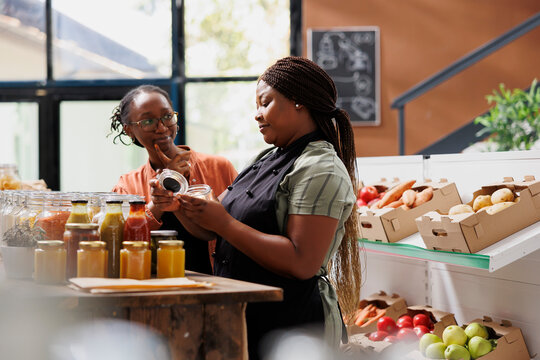 African american female vendor giving a complimentary treat to loyal customer at environmentally friendly market. Two black people talking about chemical free sustainable products at local store.
