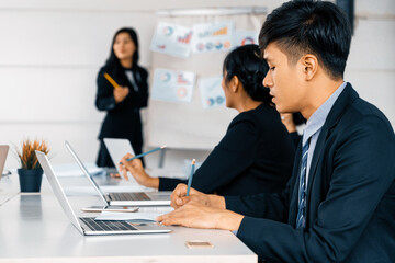 Asian business people listening to presentation from businesswoman who stands in front of group meeting room at international business conference. Multicultural corporate strategy concept. uds