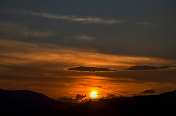 Tranquil sunset over rolling hills. The sun, low on the horizon, casts a warm, golden glow. The sky transitions from vibrant orange near the sun to deeper shades of blue as it stretches upwards