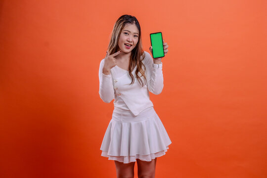 expression of beautiful teenage girl in white top standing cheerfully to camera hand holding mobile phone screen, cellphone while pointing at it isolated orange background. for technology, business