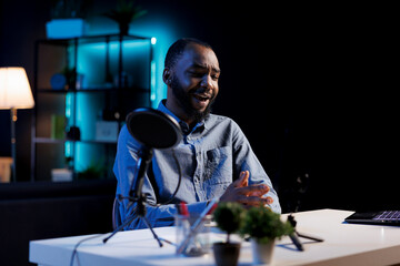 Happy online star in dark room filming reviews of various electronics, unboxing them and presenting features to viewers. Internet show host showcasing product recommendations from personal studio