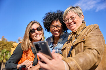 Group of diverse mature women smiling looking and using smartphone on a street. Middle-aged female...