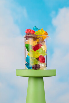 Gummy Bear Candy in Glass Beaker on chartreuse pedestal with blue sky and cloud background Happy and Joyful