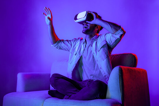 Man looking and touch to explore in simulated metaverse by using VR glasses. Happy person with headset sitting at sofa while enter in virtual world by using futuristic technology equipment. Deviation. - Powered by Adobe
