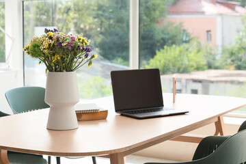 Vase with wildflowers, books and laptop on dining table in room