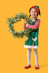 Cute little girl in elf costume with Christmas wreath on yellow background
