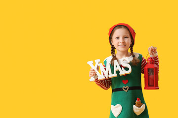 Cute little girl in elf costume with word XMAS and lantern on yellow background