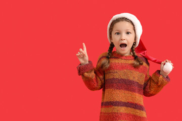 Shocked little girl in Santa hat pointing at something on red background
