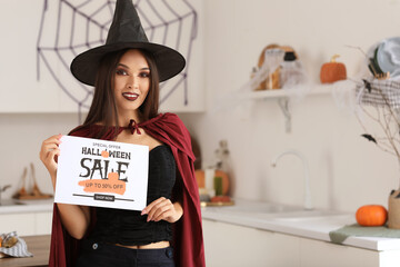 Beautiful young happy Asian woman dressed as witch with text HALLOWEEN SALE in decorated kitchen