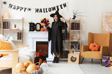 Beautiful young happy Asian woman dressed as witch for Halloween with shopping bags and gift boxes in decorated living room